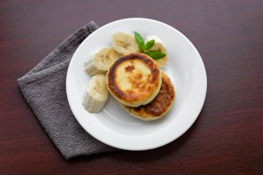 Two cottage cheese pancakes with sliced banana in a white plate. Traditional breakfast or lunch. Traditional Ukrainian breakfast.