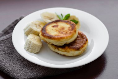 Two cottage cheese pancakes with sliced banana in a white plate. Traditional breakfast or lunch. Traditional Ukrainian breakfast.