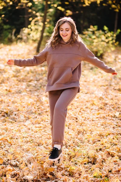 Young woman in hoodie kicks up leaves in autumn park. Sunny weather. Fall season.