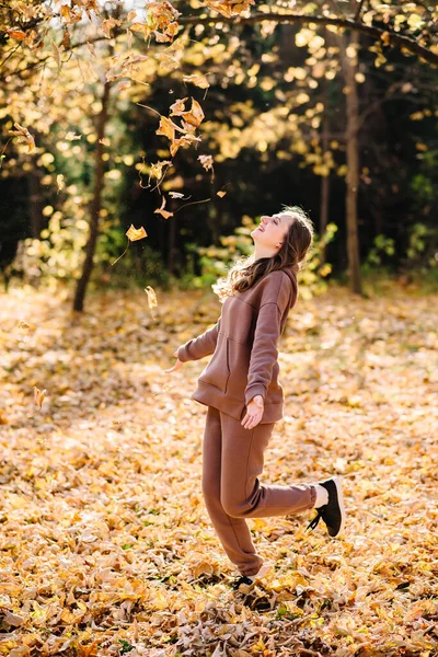 Young woman in hoodie sweater throws up leaves in autumn park. Sunny weather. Fall season.