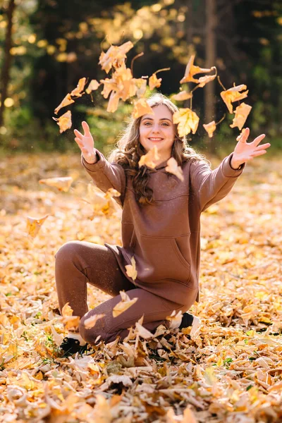 Young woman in hoodie throws up leaves in autumn park. Sunny weather. Fall season.