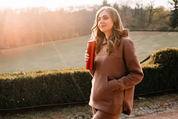 Young woman in hoodie smiling and holding a red thermo mug in an autumn park. Sunny weather. Fall season.
