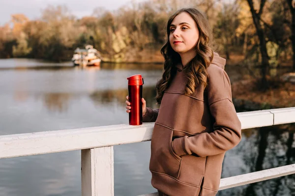 Young woman in hoodie holding a red thermo mug in an autumn park. Sunny weather. Fall season.