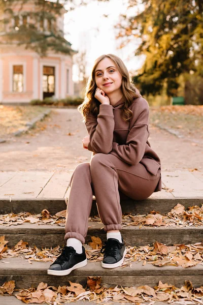 Young woman in hoodie sitting on the steps in an autumn park. Sunny weather. Fall season.