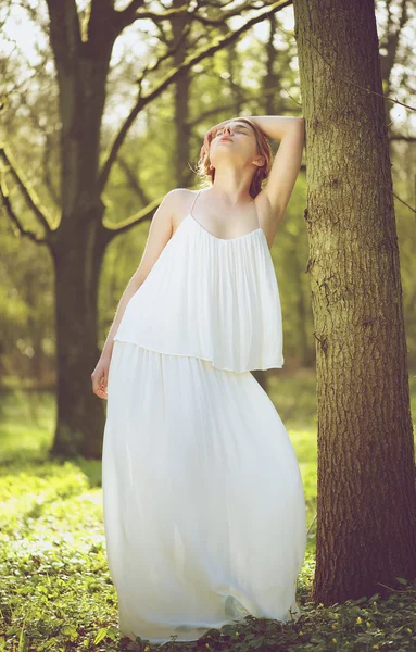 Beautiful young bride in white wedding dress posing against tree — Stock Photo, Image