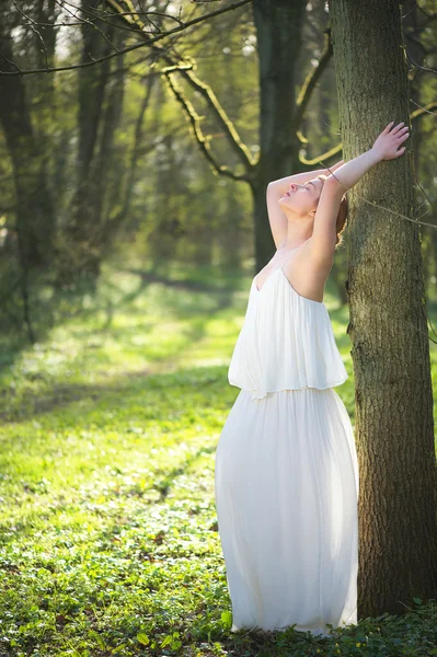 Beautiful bride in white wedding dress leaning against tree outdoors — Stock Photo, Image