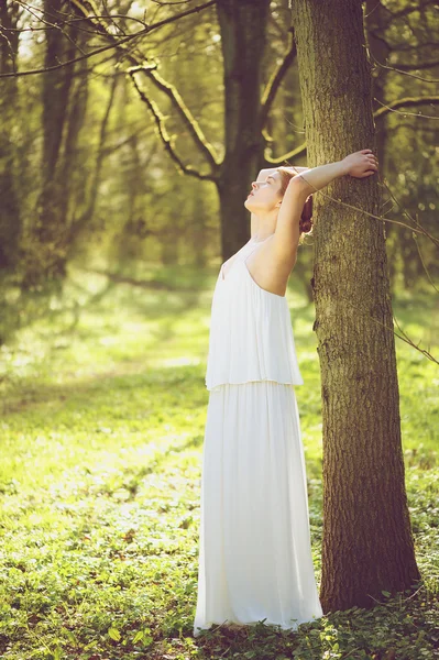 Beautiful young bride in white wedding dress leaning against tree outdoors — Stock Photo, Image