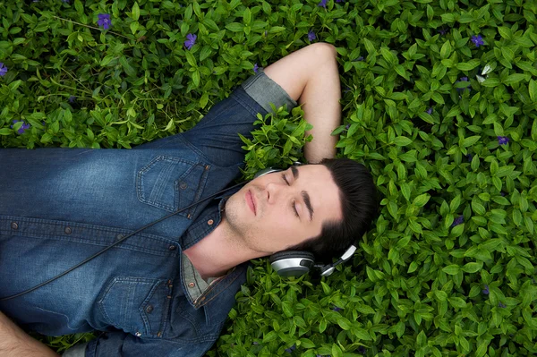 Joven descansando en la hierba con los ojos cerrados escuchando música en los auriculares — Foto de Stock