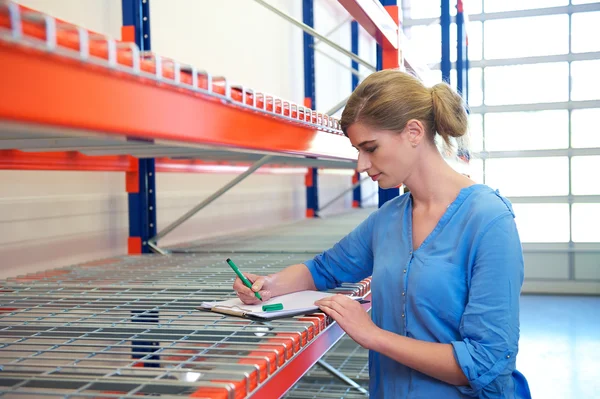 Female warehouse employee standing next to shelves and writing on clipboard — Stock Photo, Image