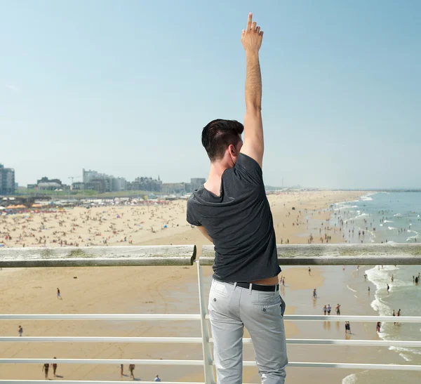 Jovem com a mão levantada apontando para cima, praia no fundo — Fotografia de Stock