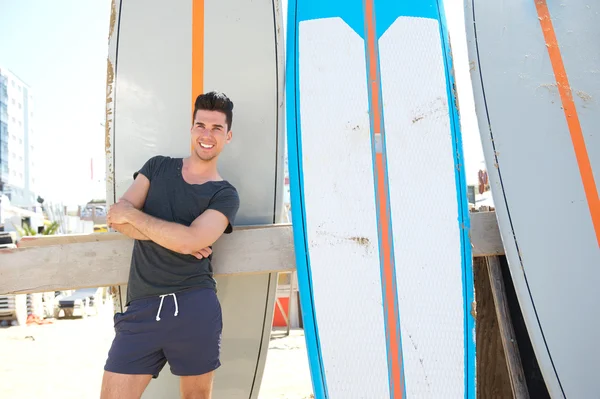 Молодий чоловік посміхається і стоїть з дошкою для серфінгу на пляжі — стокове фото