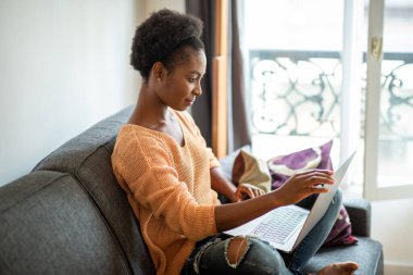Side portrait young black woman sitting on sofa at home with laptop computer
