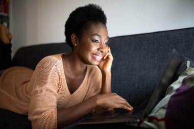 Close up portrait smiling young black woman lying on couch watching video on laptop