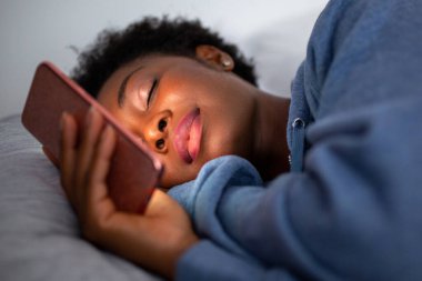 Close up portrait young African American woman sleeping in bed holding mobile phone