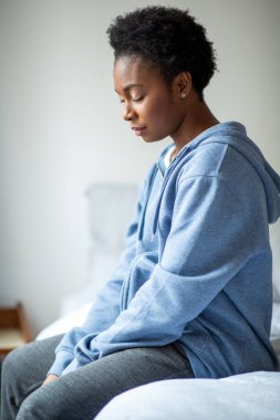 Side portrait tired young black woman sitting on bed with eyes closed