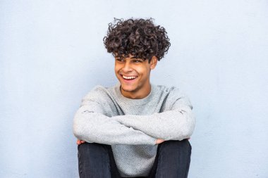 Portrait cool young guy sitting against blue wall looking away and smiling