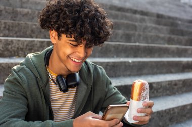 Close up portrait happy young man sitting outside with mobile phone and food in hand
