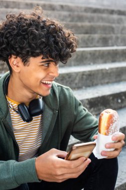 Close up portrait smiling young man sitting outside with mobile phone and food in hand
