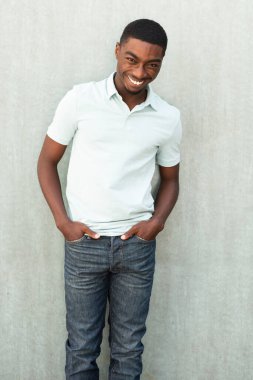 Portrait smiling young African American man standing by wall with hands in pocket