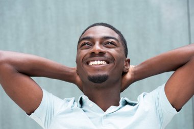 Close up portrait happy young African American man smiling with hands behind head against blue background and looking up
