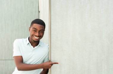 Portrait smiling young black man leaning against wall pointing finger to empty space