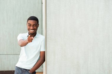Portrait handsome smiling young African American man leaning against wall and pointing finger