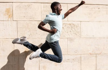 Profile portrait happy young black man jumping with arms outstretched by wall