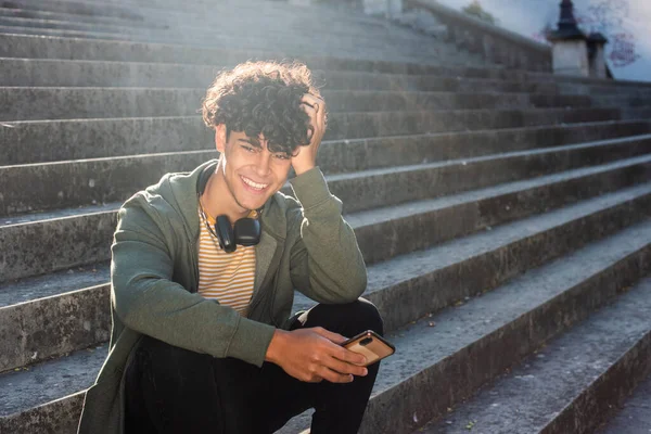 Portrait smiling young man man sitting on steps outside with mobile phone and headphones 