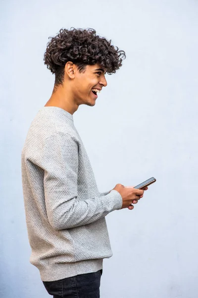 Profile portrait cool young North African man holding cellphone and laughing by gray wall