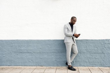 Ciddi bir Afro-Amerikan iş adamının vücut portresi cep telefonuyla duvardan duvara yaslanıyor.