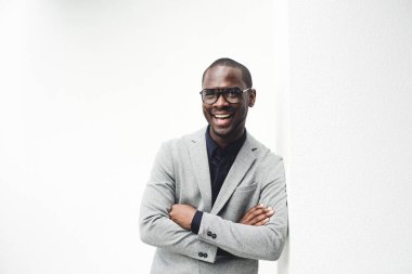 Beyaz duvara yaslanmış gözlükleriyle gülümseyen Afro-Amerikan iş adamının portresi.