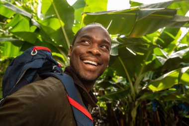 Yan taraftaki Afro-Amerikan adam ormanda sırt çantasıyla gülümsüyor. 