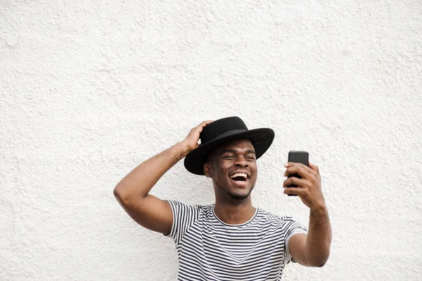 形象快乐的非洲裔美国人 拿着手机和帽子在白墙边自拍 — 图库照片