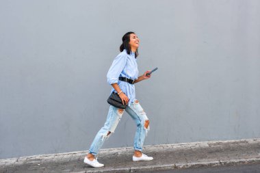 Gri duvarda cep telefonuyla yürüyen genç bir kadın vücudu.