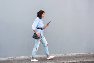 Gri duvarda cep telefonuyla yürüyen genç bir kadın vücudu.