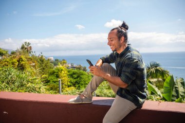 Saçı bukleli mutlu bir gezgin. Deniz manzaralı cep telefonuyla mesaj atıyor.