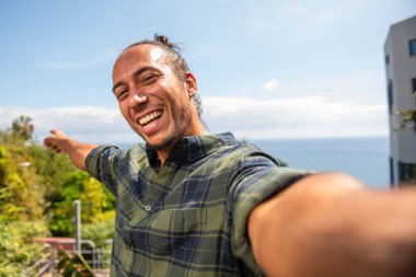 Saçlı neşeli adam selfie çekiyor ya da denize tepeden bakan blogu için yeni bir video çekiyor.