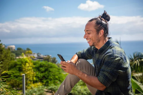 Happy man traveler with pulled up hair bun text messaging using mobile phone overlooking sea