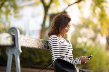 Park bankında cep telefonuyla oturan genç bir kadının yan portresi.