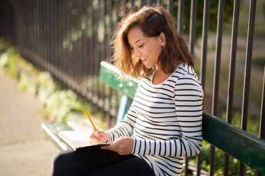 Bankta oturup kitap yazan genç bir kadının yan portresi.