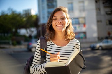 Şehirde kitap ve çantayla gülümseyen kız üniversite öğrencisinin portresi.