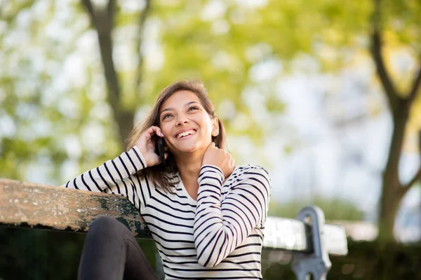 Mutlu genç bir kadının portresi bankta oturuyor ve cep telefonuyla konuşuyor.