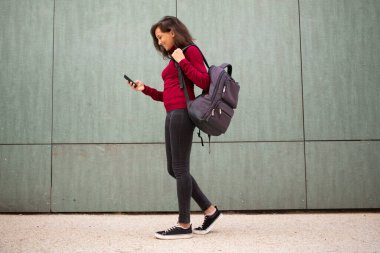 Duvarda cep telefonu ve çantasıyla yürüyen genç bir kadının tam vücut profili.