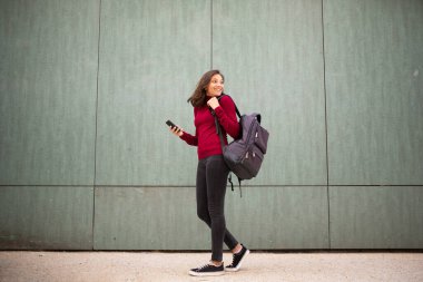 Duvardan duvara çantalı ve cep telefonlu genç bir kadının tam boy portresi.