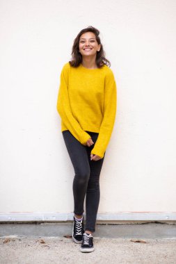 Beyaz duvara yaslanmış gülümseyen genç bir kadının vücut portresi.