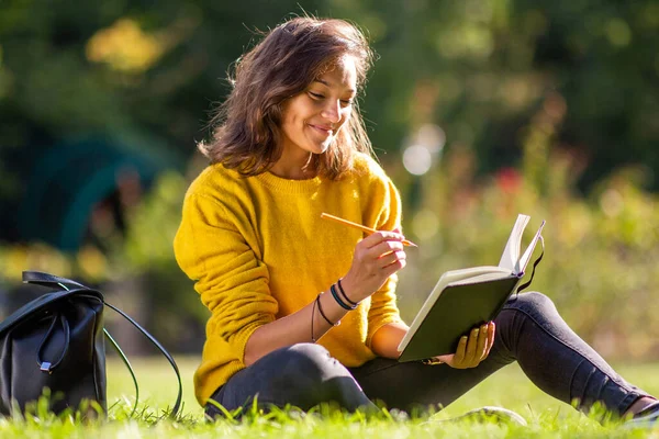 本の中で書く公園の草の上に座って笑顔の若い女性の肖像 — ストック写真