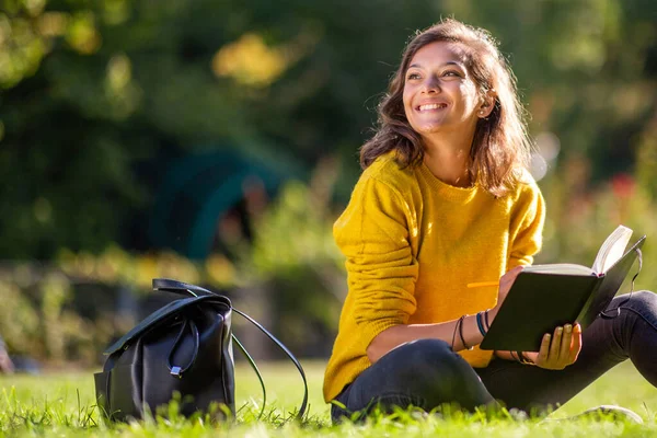 公園で草の上に座って本の中で書くと離れて見て幸せな若い女性の肖像画 — ストック写真