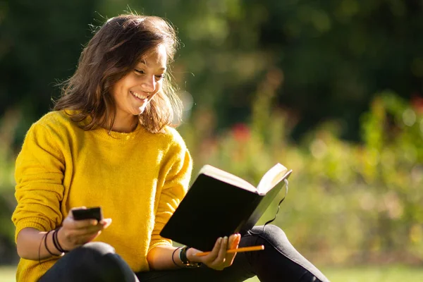 本や携帯電話と公園で草の上に座って幸せな若い女性の肖像画 — ストック写真