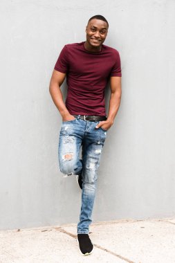 Gri duvara yaslanmış ve gülümseyen yakışıklı Afro-Amerikan bir adamın tam boy portresi.
