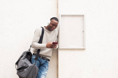 Çantalı ve kulaklıklı beyaz duvarın yanında dururken cep telefonu kullanan Afrikalı Amerikalı bir adamın portresi. 